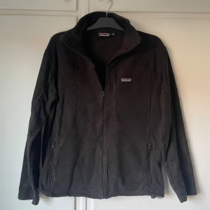 Svart fleece tröja/jacka från Patagonia M - Mysig svart fleecejacka från Patagonia med hel dragkedja framtill och två sidofickor med dragkedja. Klassisk ståkrage och broderad logga på bröstet. Perfekt för kyliga dagar och enkel att slänga på sig över en hoodie.