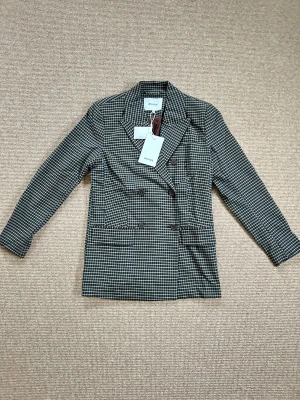 Rutig kavaj från Minus med knappar - Snygg rutig kavaj från Minus i brunt och ljusblått med dubbelknäppning och klassisk krage. Kavajen har långa ärmar, två fickor framtill och är fodrad. Perfekt för dig som vill ha en stilren och trendig look. 
