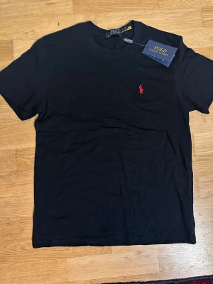 Svart t-shirt från Polo Ralph Lauren - Svart t-shirt från Polo Ralph Lauren med klassisk röd broderad logga på bröstet. T-shirten har rund halsringning och korta ärmar. Tillverkad i mjuk bomull för en skön känsla och stilren look.