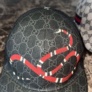 Gucci keps med orm och GG-mönster - Snygg keps från Gucci med klassiskt GG-monogram i mörkgrått och svart. Framsidan pryds av en röd och vit orm, och baksidan har mesh och justerbart band med röd/blå rand. Perfekt statement-piece för dig som gillar lyxiga accessoarer. Varan är Autentisk och har kvitto! Snabb frakt!   Pris kan diskuteras vid snabb affär!