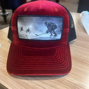 Röd keps med hockeymotiv - Snygg röd keps i sammet och polyester med svart mesh på sidorna och bak. Framtill finns ett stort svartvitt tryck med hockeyspelare i action. Justerbar passform med plastspänne. Perfekt för dig som gillar sportig stil och hockey.