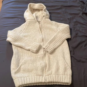 Beige stickad hoodie med dragkedja - Mysig beige stickad hoodie med huva och dragkedja framtill. Tröjan har långa ärmar med breda ribbade muddar och två praktiska fickor på framsidan. Perfekt för att hålla sig varm under kyliga dagar.