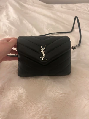 Svart quiltad axelväska från YSL - Snygg svart axelväska från YSL med quiltad design och silverfärgat YSL-logo framtill. Väskan har en stilren form och är tillverkad i skinn med justerbar axelrem. Perfekt för dig som vill ha en lyxig och trendig accessoar.INGEN BESTÄLLNINGSVARA