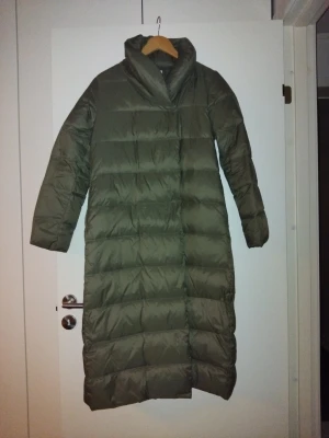 Lång grön omlott dunjacka med hög krage - Säljer en lång, olivgrön dunjacka med hög krage och quiltad design. Jackan har en rak passform och långa ärmar, perfekt för kalla dagar. Den är stilren och enkel utan synliga detaljer som knappar eller dragkedja framtill.