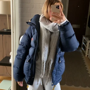Blå dunjacka från Fjällräven - Säljer en blå dunjacka från Fjällräven med huva, gula snören och dragkedja samt knappar framtill. Jackan har två stora fickor med lock och klassisk Fjällräven-logga på bröstet. Perfekt för kalla dagar och riktigt snygg passform. Nypris 7,499. Har haft den sen förra året men inte kommit till användning. Inga skador eller fläckar alls!
