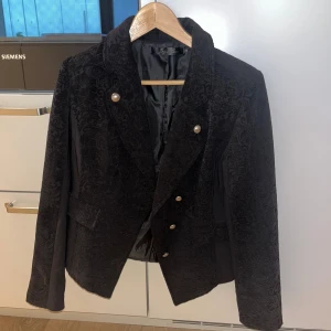 Svart sammet kavaj med pärlknappar - Snygg svart kavaj i sammet med diskret mönster och pärlknappar framtill. Klassisk krage och figurnära passform, perfekt för att lyfta vilken outfit som helst. Långa ärmar och elegant look.