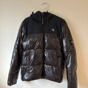 Calvin Klein - Pufferjacka - En svart pufferjacka från Calvin Klein med glansig finish och huva. Jackan har dragkedja framtill, två stora fickor och diskret CK-logga på bröstet och ärmen. .