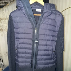 Mörkblå Moncler cardigan  - Snygg mörkblå cardigan från Moncler med stickade ärmar och rygg. Jackan har vadderad framsida, dragkedja, två fickor med tryckknappar och en huva. Perfekt för dig som vill ha en stilren och exklusiv look under kyliga dagar.