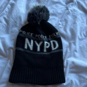 Svart NYPD mössa med tofs - Säljer en svart och grå stickad NYPD-mössa med stor tofs på toppen. Mössan har texten 'POLICE' och 'NYPD' i vitt samt ett broderat NYPD-märke framtill. Ribbad kant och klassisk streetstil, perfekt för kalla dagar.