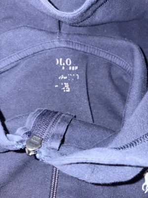 Mörkblå hoodie från Polo Ralph Lauren - Snygg mörkblå hoodie från Polo Ralph. Väldigt skön men har dock några små tecken på användning. En på sista bilden att texten har försvunnit och vid för man spänner åt den att det inte finns något snörre den gick sönder och tog bort den. Nypris 2000kr