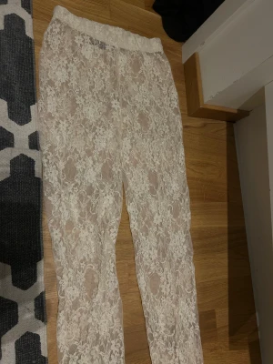 Vita spetsbyxor med blommönster - Säljer ett par vita byxor i transparent spets med blommigt mönster. De har elastisk midja och är raka i modellen, perfekta för dig som vill sticka ut med en unik look. Materialet är mjukt och luftigt, passar dig som gillar detaljer och romantisk stil.