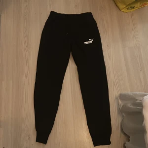 Svarta mjukisbyxor från Puma - Svarta mjukisbyxor från Puma med vit logga på vänster lår. Byxorna har ribbade muddar vid bensluten och elastisk midja med snörning. Perfekta för chill eller träning, riktigt sköna och stilrena.