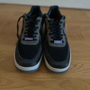 Nike Air Force 1 svart/grå sneakers - Säljer ett par Nike Air Force 1 sneakers i svart och grått med vit sula. Skorna har klassisk snörning, rund tå och detaljer i både skinn och mocka. Swoosh-loggan i grått på sidan och coola färgdetaljer på snörningen. Perfekt för dig som gillar streetstyle.