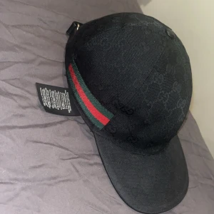 Svart gucci keps helt ny skick gamla modellen/black label  - Snygg svart keps från Gucci med diskret GG-monogram över hela kepsen. På sidan finns en klassisk röd och grön rand som ger den extra lyxig vibe. Justerbar baktill med metallspänne. Perfekt för dig som vill ha en stilren och exklusiv look.