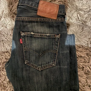 Levi's 501 jeans mörkblå 30/32 - Säljer ett par klassiska Levi's 501 jeans i mörkblå tvätt med snygga slitningar. Modellen har raka ben, fem fickor och den ikoniska läderpatchen bak i midjan. Jeansen har orange kontrastsömmar och är tillverkade i robust denim.