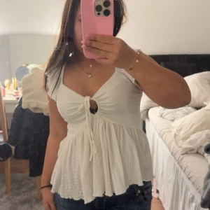 Vit omlott blus med knyt och volang - Supersöt vit blus med omlott och knyt framtill, v-ringad och med volang vid axlarna. Blusen har ett lätt rynkat tyg och är ärmlös, med en flowy passform som ger en chill vibe. Perfekt till jeans eller kjol för en fräsch look.