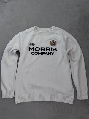 Vit stickad tröja från Morris Company - Snygg vit stickad tröja från Morris Company med broderad logga och text framtill. Tröjan har rund hals, långa ärmar och ribbade muddar vid ärmslut och nederkant. Perfekt för dig som gillar klassisk stil med en modern touch.