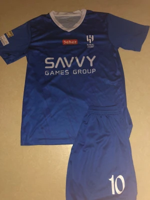 Blå Al Hilal Neymar Jr fotbollströja - Säljer en blå Al Hilal fotbollströja med Neymar Jr och nummer 10 på ryggen. Tröjan har vit V-ringad krage, korta ärmar och tryck från Savvy Games Group och Jahez. Materialet är lätt och andas, perfekt för match eller träning. Matchande blå shorts med nummer 10 ingår.
