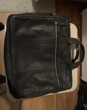 Svart tiger of sweden portfölj i skinn med dragkedja - Stilren svart portfölj i äkta skinn med praktiskt handtag och en snygg dragkedjedetalj på framsidan. Perfekt för att bära laptop, papper och andra viktiga grejer till skolan eller jobbet. Enkel och klassisk design som passar till allt. Ny pris 3000kr.