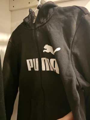 Svart hoodie från Puma med dragkedja - Svart hoodie från Puma i storlek M med vit logga framtill. Tröjan har huva, hel dragkedja och två fickor. Materialet är mjuk bomull och polyester, perfekt för chill eller träning. Klassisk streetwear-look med bekväm passform och lång ärm.