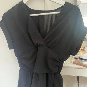 Svart omlotttopp med v-ringning - Svart topp med snygg v-ringning och omlott-detalj framtill. Toppen har korta ärmar och ett smockat parti i midjan som ger en skön passform. Materialet är lätt och luftigt, perfekt för varmare dagar. Stilren och enkel design som är lätt att matcha.