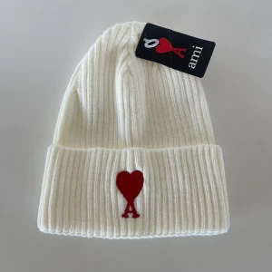 Vit ribbad mössa A - Vit ribbstickad mössa från A med uppvikt kant och röd broderad logga med hjärta och bokstaven A framtill. Snygg och enkel design som ger en clean look, perfekt för att lyfta din outfit. Mjuk och varm, tillverkad i ull.