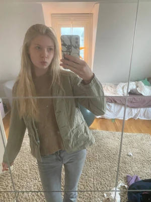 Olivgrön jeansjacka - Olivgrön jeansjacka med snygg broderad blomdetalj på ena fickan, syns dock ej jättemycket från långt håll. Jackan har knappar framtill och två fickor. Perfekt för dig som gillar en chill och Stockholms stil