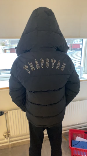 Trapstar svart pufferjacka med huva - Säljer en svart pufferjacka från Trapstar med stor huva och feta broderade bokstäver på ryggen. Jackan har dragkedja framtill, två stora fickor och en rund Trapstar-logga på bröstet. Perfekt för dig som vill ha streetwear-vibe och hålla dig varm. Den är helt ny, jag har använt den 2 gånger och jag har kvar taggen.