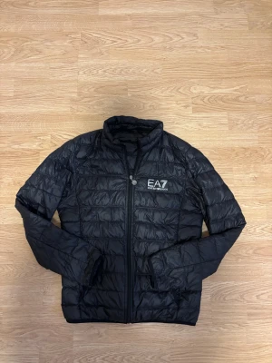 Svart pufferjacka EA7 Emporio Armani - Svart pufferjacka från EA7 Emporio Armani med quiltad design och hög krage. Jackan har dragkedja framtill, två sidofickor och EA7-logga på bröstet. Perfekt för dig som gillar stilren och sportig look.