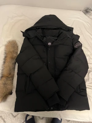 Canada Goose - Säljer en svart dunjacka från Canada Goose med huva och avtagbar pälskant. Jackan har quiltad design, lång ärm och klassisk logotyp på ärmen. Perfekt för kalla vinterdagar och har både dragkedja och knappar framtill.