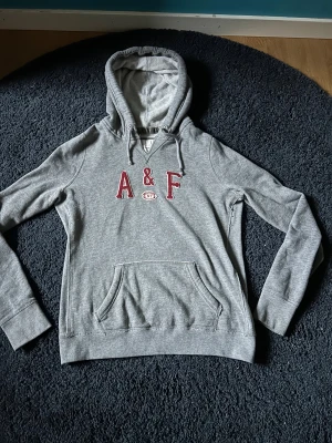 Grå hoodie Abercrombie & Fitch - Snygg grå hoodie från Abercrombie & Fitch med broderad A&F-logga i vinrött framtill. Klassisk känguruficka, dragsko i huvan och mjukt bomullsmaterial. Perfekt för en chill och avslappnad stil.