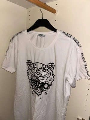 Vit t-shirt med tigertryck - Snygg vit t-shirt från Kenzo med ikoniskt tigertryck i svart på bröstet och glansiga detaljer längs axlarna där Kenzo-loggan syns. Rund halsringning och korta ärmar. Materialet är mjuk bomull som känns skönt mot huden.