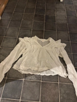 Vit blus med volang från Gina Tricot - Supersöt vit blus från Gina Tricot med volangdetaljer på axlarna och v-ringning. Blusen har långa, lätt transparenta ärmar med smock vid ärmslut och spetsinslag över bröstet. Luftigt material som ger en romantisk vibe. Gått sönder lite därbak men de går att fixa 