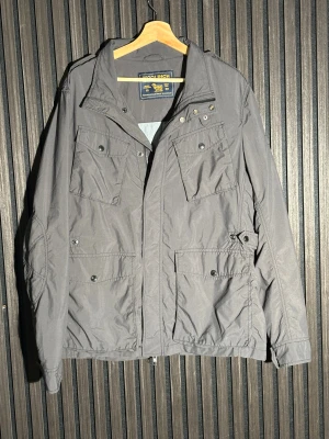 Grå vindjacka från Woolrich - Säljer en grå vindjacka från Woolrich med fyra stora fickor framtill, hög krage och snygga detaljer vid axlarna. Jackan har dragkedja och tryckknappar, samt diskret logga på ärmen. Perfekt för dig som gillar stilren och funktionell design. Priset är ej hugget i sten. Nypris 6000kr.