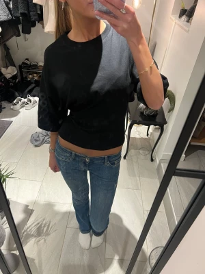 Svart omlottopp med knyt i ryggen - Svart topp med rund hals och trekvartsärm. Toppen har en croppad passform och snygg omlottdetalj med knyt i ryggen som ger en öppen rygg. Perfekt för dig som gillar stilrena plagg med en twist.