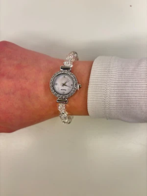 Glittrig klocka med pärlarmband - Superfin klocka med glittrande stenar runt urtavlan och ett armband av genomskinliga pärlor. Urtavlan är vit med diskreta markeringar och silverdetaljer. Perfekt accessoar för dig som gillar bling och vill sticka ut lite extra.