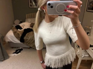 Vit stickad topp med fransar från H&M - Supersnygg vit stickad topp från H&M med korta ärmar och rund hals. Nederdelen har coola fransdetaljer som ger en unik look. Perfekt att styla med jeans eller skinnbyxor för en trendig vibe. Materialet är mjukt och bekvämt stickat.