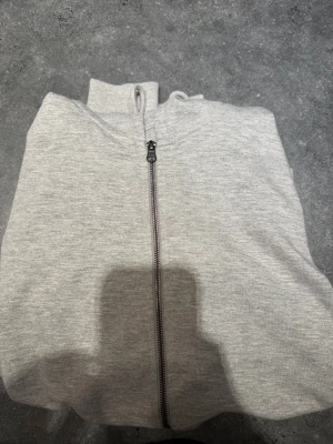 Grå hoodie med dragkedja Jack & Jones - Säljer en stilren ljusgrå hoodie från Jack & Jones Essentials i storlek S. Tröjan har hel dragkedja framtill, huva med snörning och ribbade muddar. Aldrig använd.