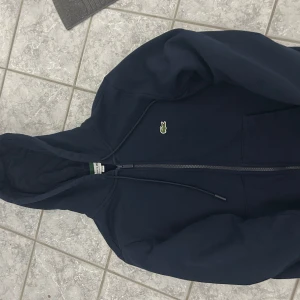 Marinblå hoodie från Lacoste - Snygg marinblå hoodie från Lacoste med klassisk krokodillogga på bröstet. Tröjan har huva med snörning, dragkedja framtill och rymliga fickor. Tillverkad i mjuk bomull och har en normal passform som är chill och enkel att bära.