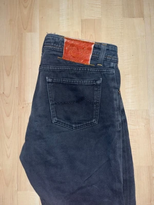 Jacob Cohen svarta jeans, storlek 33 - Svarta jeans från Jacob Cohen, tillverkade i Italien. Klassisk femficksmodell med orangea detaljer och logotyp på bakfickan. Jeansen har rak passform och är gjorda i mjukt bomullsmaterial med knappgylf. Snyggt läderpatch baktill.