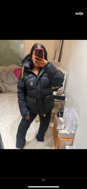 Svart dunjacka från Fjällräven - Säljer en svart, puffig dunjacka från Fjällräven med huva och gula snören. Jackan har dragkedja och knappar framtill, två fickor med lock och Fjällräven-logga på bröstet. Perfekt för kalla dagar och har en cool, oversized vibe.