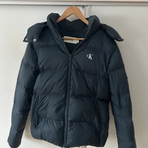 Svart pufferjacka från Calvin Klein - Svart pufferjacka från Calvin Klein med CK-logga på bröstet. Jackan har huva, dragkedja framtill och två sidofickor med dragkedja. Jackan är i small men passar även mig som är m