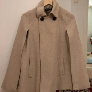 Beige cape/kappa från Zara med knappar - Snygg beige kappa/cape från Zara med klassisk krage och två svarta knappar framtill. Jackan har en A-linjeformad siluett, diskreta fickor och är tillverkad i ett ulligt material som känns mjukt. Perfekt för dig som gillar stilrena och tidlösa plagg.
