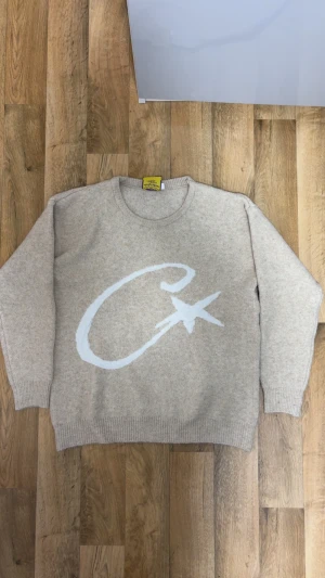 Cortez stickad tröja  - Säljer en beige stickad tröja från Corteiz med ett stort vitt tryck av bokstaven C och en stjärna på framsidan. Tröjan har rund hals, ribbade muddar och lång ärm. Perfekt för dig som gillar chill och snygga detaljer.