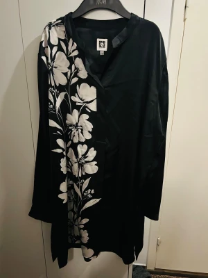 Svart blommig kortklänning Anne Klein - Svart kortklänning från Anne Klein med lång ärm och snyggt blommigt mönster i vitt på ena sidan. Klänningen har en loose passform och är tillverkad i ett glansigt, mjukt polyestermaterial som känns lyxigt mot huden. Perfekt för dig som gillar stilrena kontraster och unika detaljer.