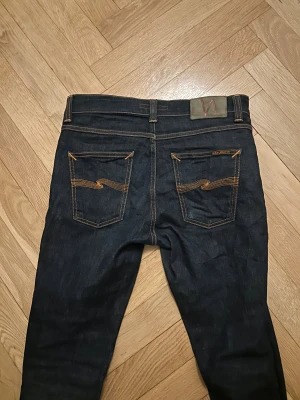 Mörkblå Nudie Jeans Skinny W29 L32 - Säljer ett par mörkblå skinny jeans från Nudie Jeans med klassiska orange sömmar och snygga broderade bakfickor. Modellen har smal passform och raka ben, tillverkade i slitstarkt jeanstyg. Perfekt för dig som gillar stilrena och moderna jeans. Köpt helt nästan helt nya. Passar perfekt för dig som behöver ett par nya jeans. Säljer bara för att jag har växt ur dem har bara haft på dem få tal gånger, inga fläckar eller något liknande. Pris kan diskuteras 