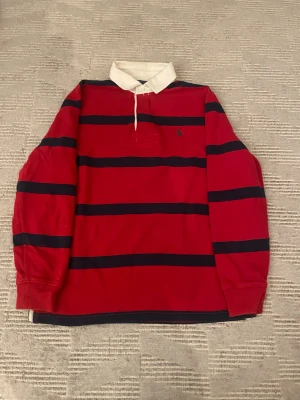 Röd och marinblå randig pikétröja Polo Ralph Lauren - Snygg pikétröja från Polo Ralph Lauren i rött med breda marinblå ränder och klassisk vit krage. Tröjan har långärmad passform, knappar vid halsen och broderad logga på bröstet. Tillverkad i mjuk bomull, perfekt för en chill och sportig stil.