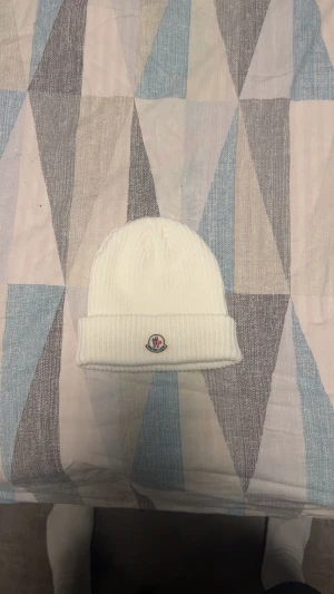 Vit ribbstickad mössa från Moncler - Vit ribbstickad mössa från Moncler med klassisk uppvikt kant och broderad logotyp framtill. Tillverkad i mjukt material som håller dig varm och snygg under kalla dagar. Perfekt accessoar för dig som gillar stilrena och exklusiva detaljer.
