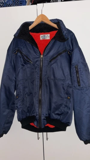 Blå bomberjacka Fristads XL med fleece - Säljer en mörkblå bomberjacka från Fristads i storlek XL. Jackan har röd och svart fleecefoder, flera dragkedjefickor och ribbade muddar. Perfekt för kalla dagar med sin varma insida och coola look.