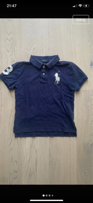 Marinblå piké från Polo Ralph Lauren - Säljer en klassisk marinblå pikétröja från Polo Ralph Lauren med vit broderad logga på bröstet och en stor vit siffra 3 på ärmen. Tröjan har krage, korta ärmar och knappar framtill. Materialet är mjuk bomull som andas och känns skön mot huden. original storlek m barn men tvättad till s